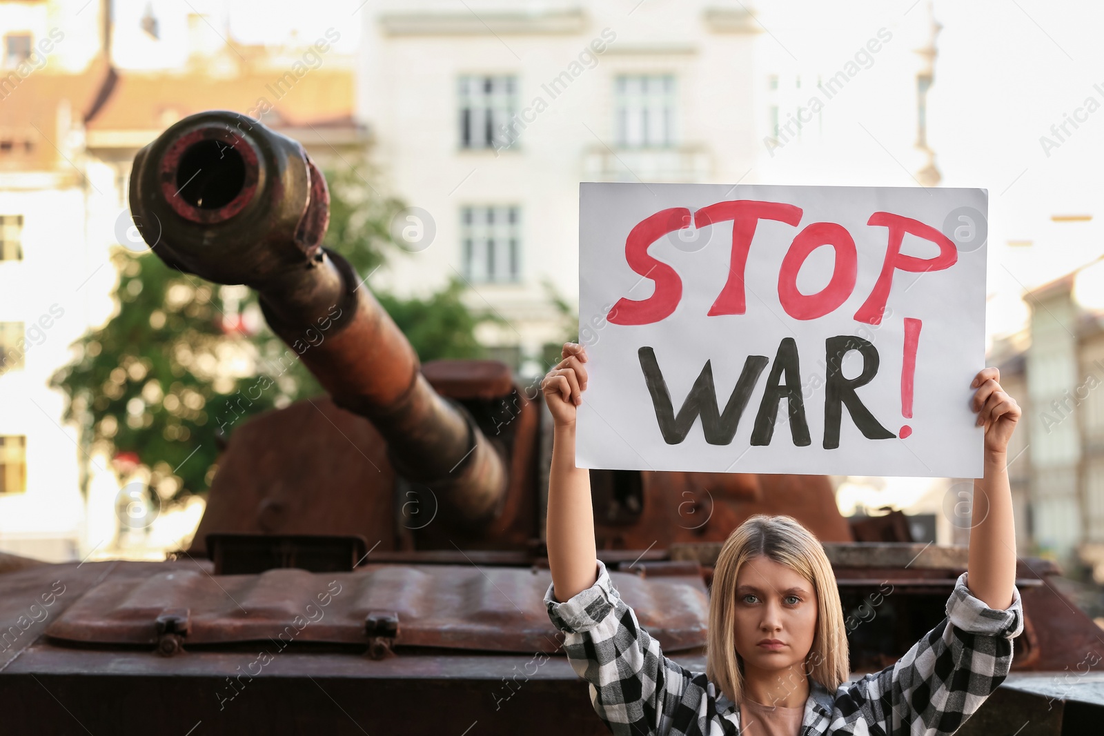 Photo of Sad woman holding poster with words Stop War near broken tank in city