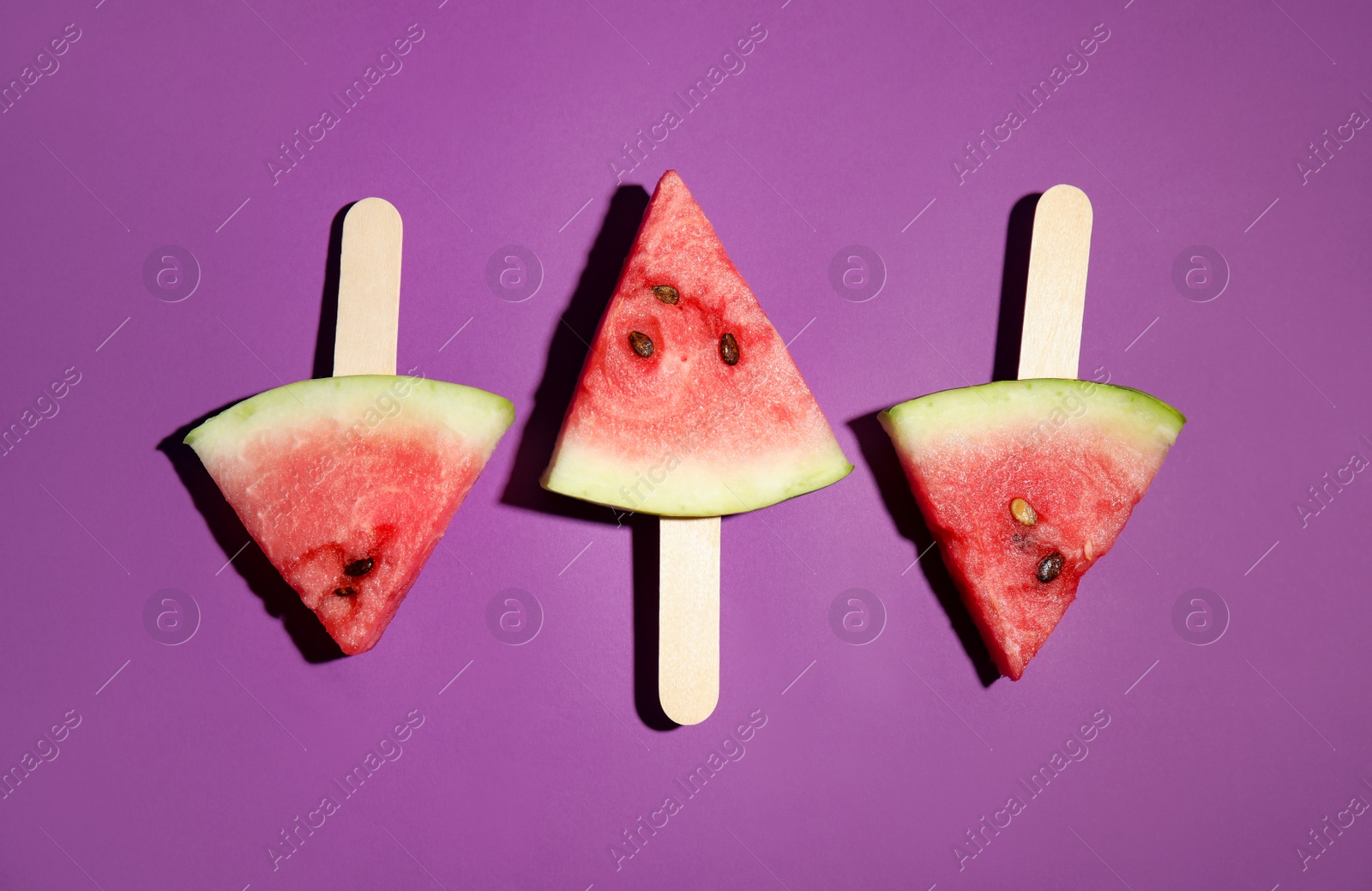 Photo of Watermelon slices with wooden sticks on purple background, flat lay