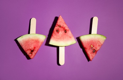 Photo of Watermelon slices with wooden sticks on purple background, flat lay