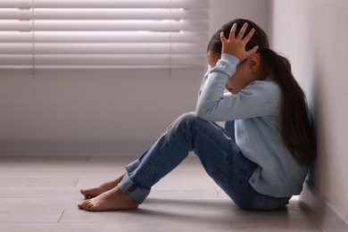 Photo of Child abuse. Upset little girl sitting on floor near light wall indoors