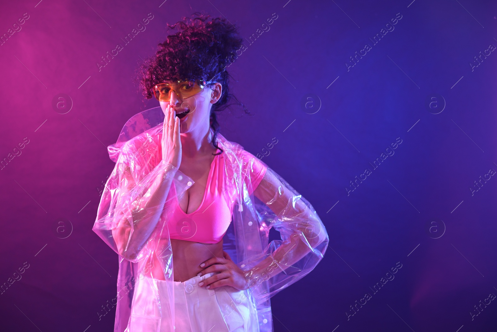 Photo of Beautiful young woman in transparent coat and sunglasses posing on color background in neon lights. Space for text