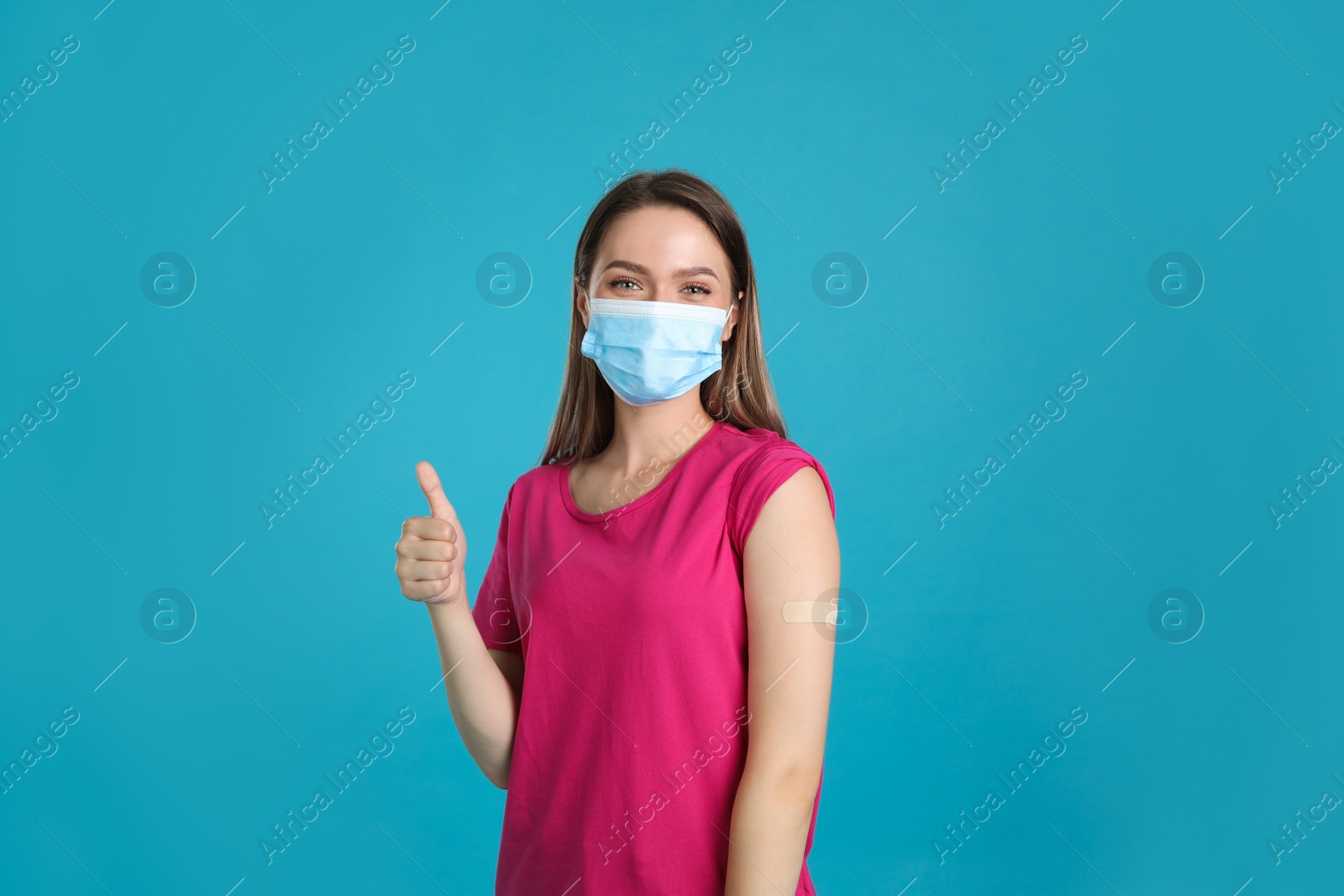 Photo of Vaccinated woman with protective mask and medical plaster on her arm showing thumb up against light blue background