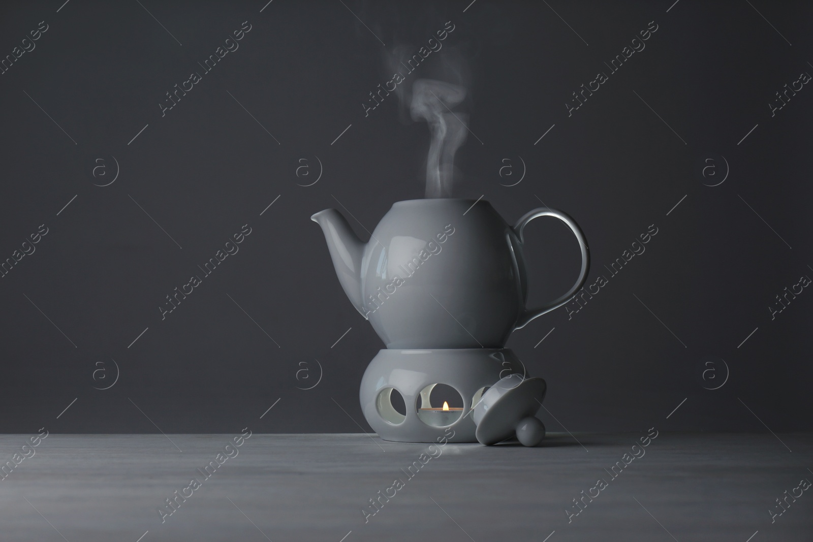 Photo of Ceramic teapot and warmer with burning candle on table against grey background