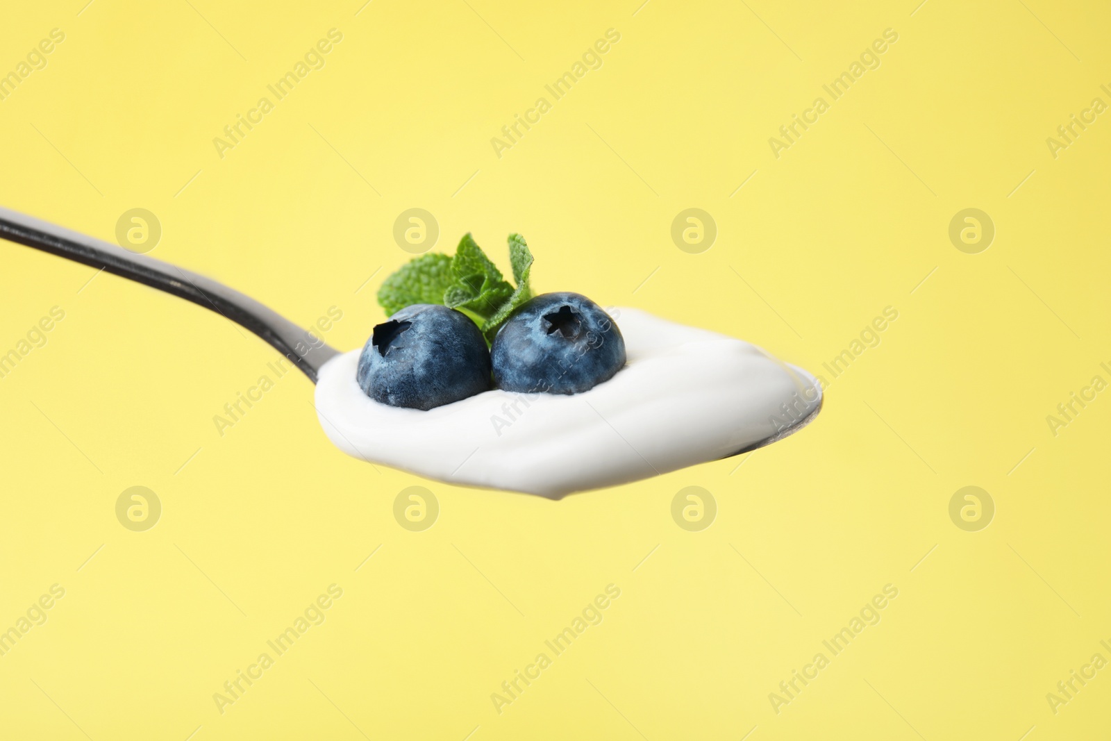 Photo of Spoon with yogurt, blueberries and mint on yellow background, closeup
