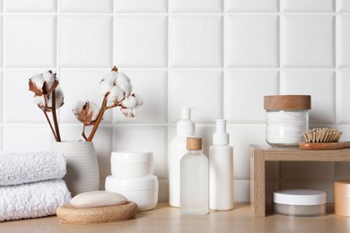 Different bath accessories, personal care products and cotton flowers in vase on wooden table near white tiled wall