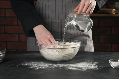 Photo of Making bread. Woman pouring water into bowl with flour at grey textured table in kitchen, closeup