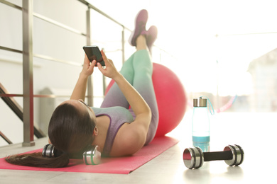 Photo of Lazy young woman with smartphone on yoga mat indoors