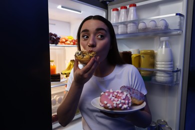 Photo of Young woman eating donut near refrigerator in kitchen at night. Bad habit