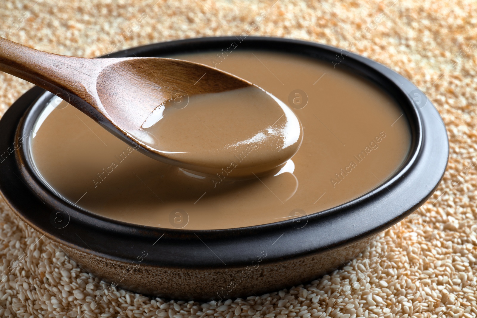 Photo of Spoon of tasty sesame paste above bowl on seeds, closeup