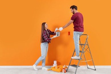 Photo of Man taking can of paint from woman near orange wall indoors. Interior design
