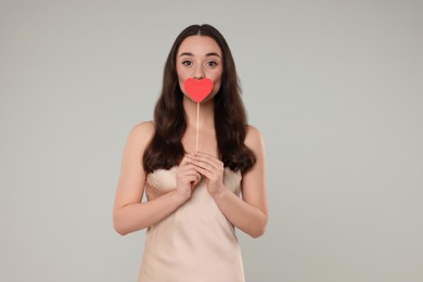 Photo of Beautiful young woman covering her mouth with paper heart on grey background