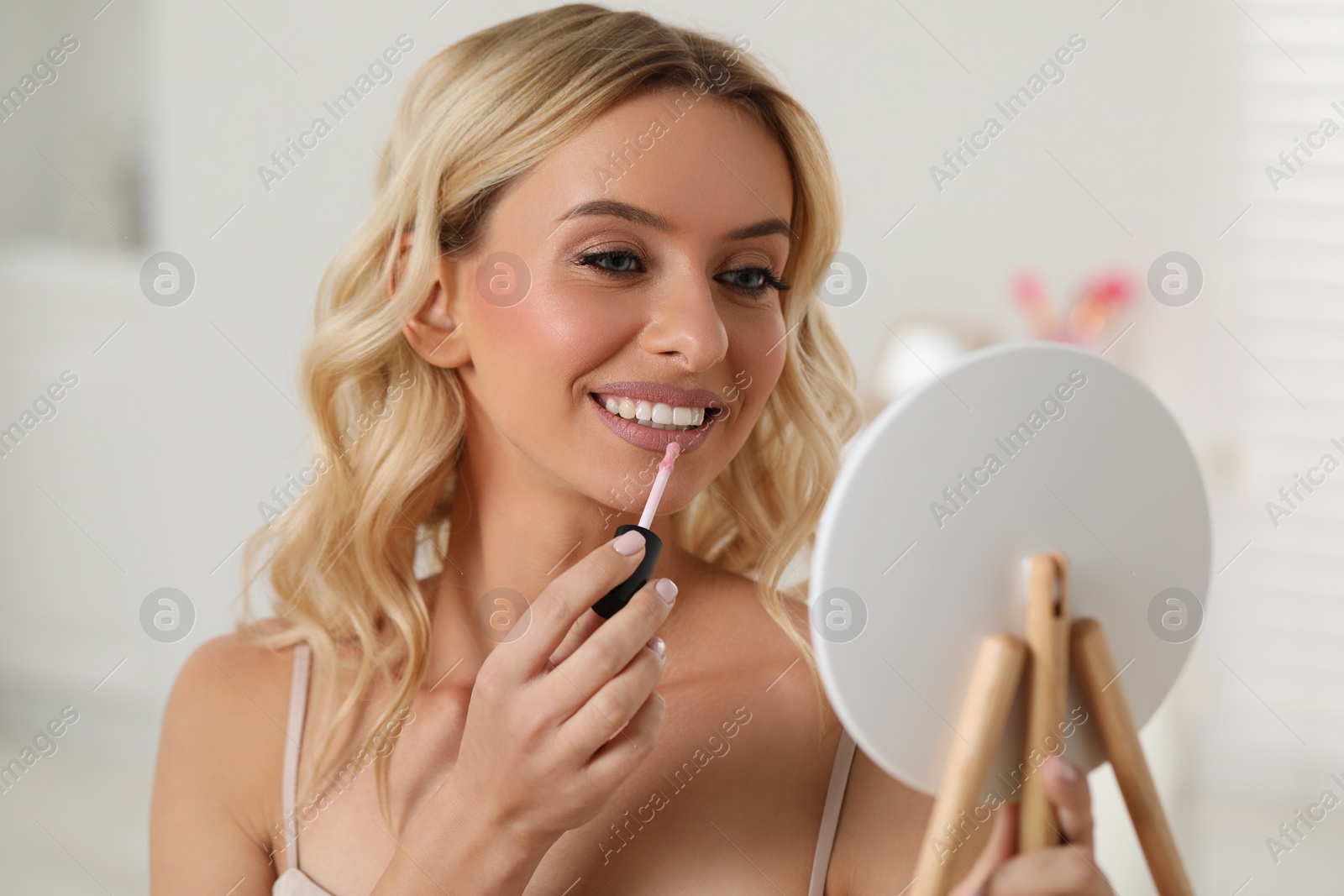 Photo of Beautiful makeup. Smiling woman with mirror applying liquid lipstick indoors