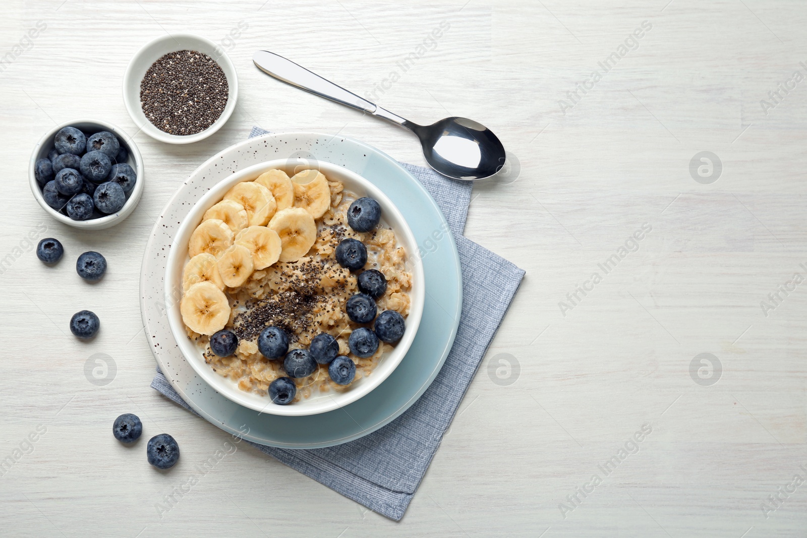 Photo of Tasty oatmeal with banana, blueberries and chia seeds served in bowl on white wooden table, flat lay. Space for text