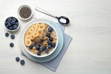 Tasty oatmeal with banana, blueberries and chia seeds served in bowl on white wooden table, flat lay. Space for text