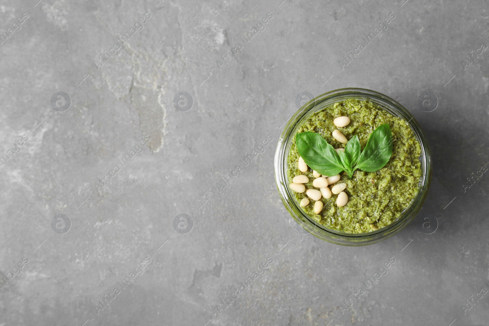 Photo of Jar with delicious pesto sauce, pine nuts and basil leaves on grey table, top view. Space for text