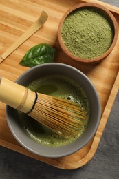 Photo of Cup of fresh matcha tea with bamboo whisk, green powder and spoon on dark grey table, top view