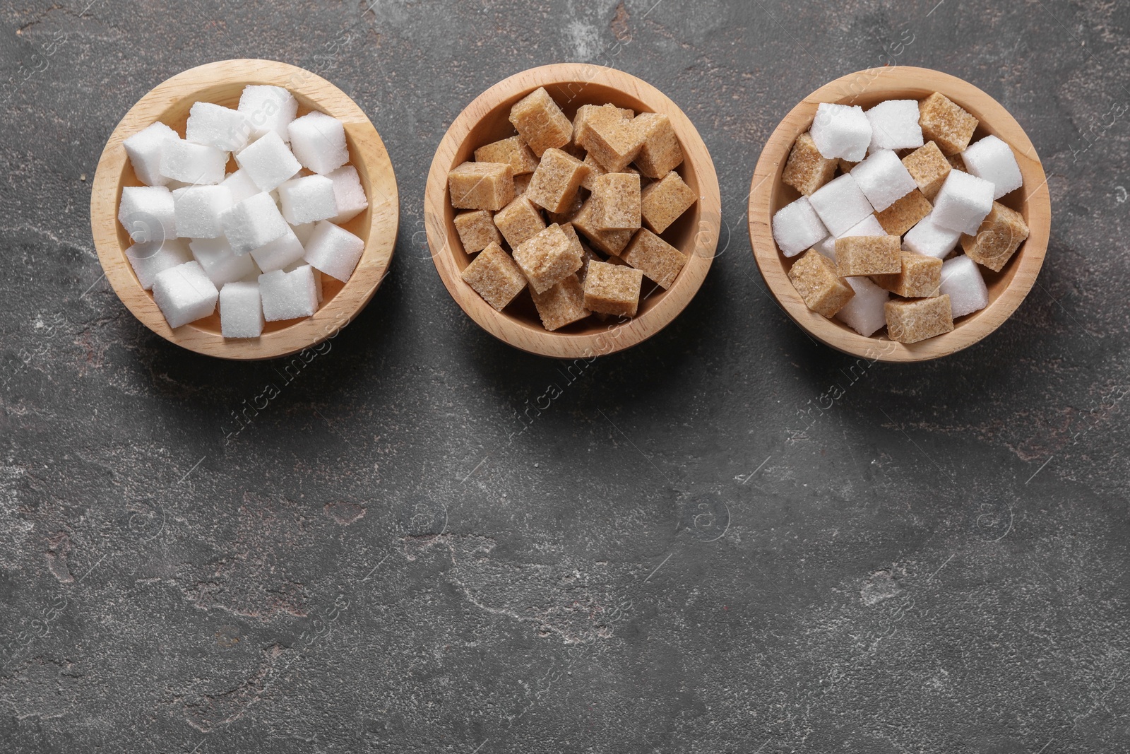 Photo of Different sugar cubes in bowls on gray textured table, flat lay. Space for text