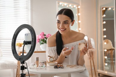 Photo of Beauty blogger with makeup product recording video in dressing room at home. Using ring lamp and smartphone