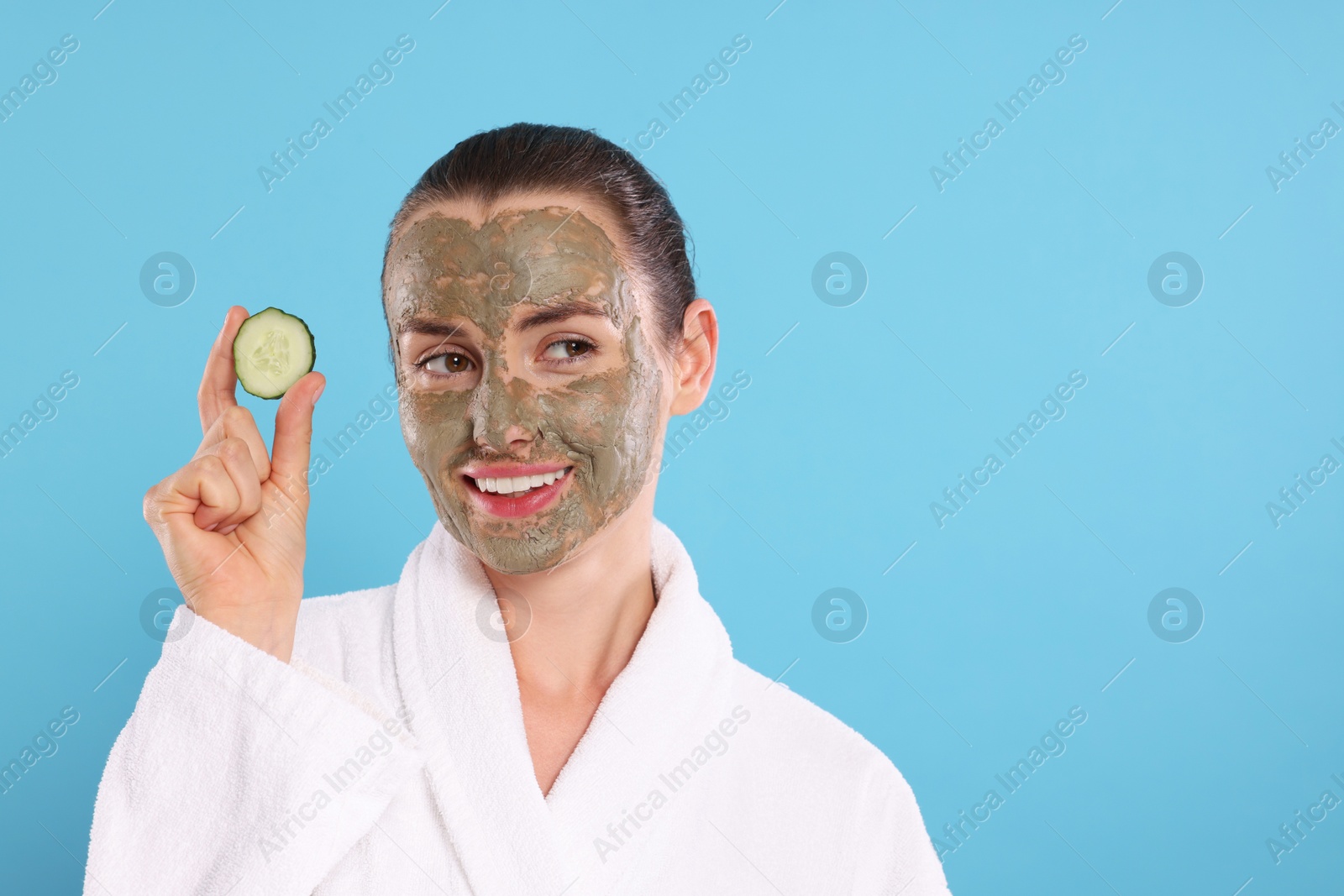 Photo of Beautiful woman with clay mask holding piece of cucumber on light blue background, space for text