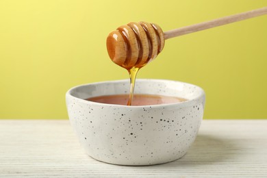 Photo of Pouring tasty honey from dipper into bowl at white wooden table against yellow background, closeup