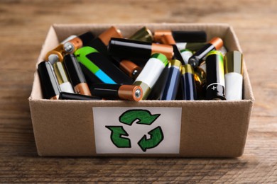 Used batteries in cardboard box with recycling symbol on wooden table, closeup