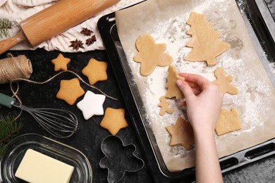 Photo of Woman putting raw Christmas cookies on baking tray at dark table, top view