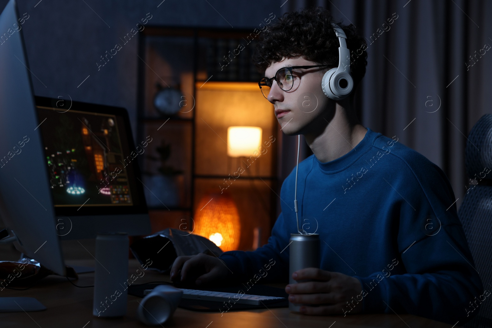 Photo of Young man with energy drink and headphones playing video game at wooden desk indoors