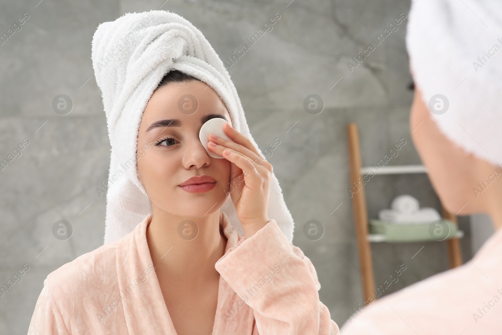 Photo of Beautiful woman in terry towel removing makeup with cotton pad near mirror indoors