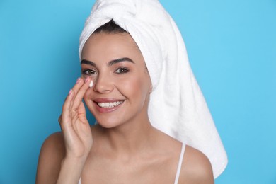 Beautiful young woman with towel applying cream on face against light blue background