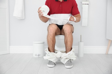 Photo of Young man with bath tissue sitting on toilet bowl at home