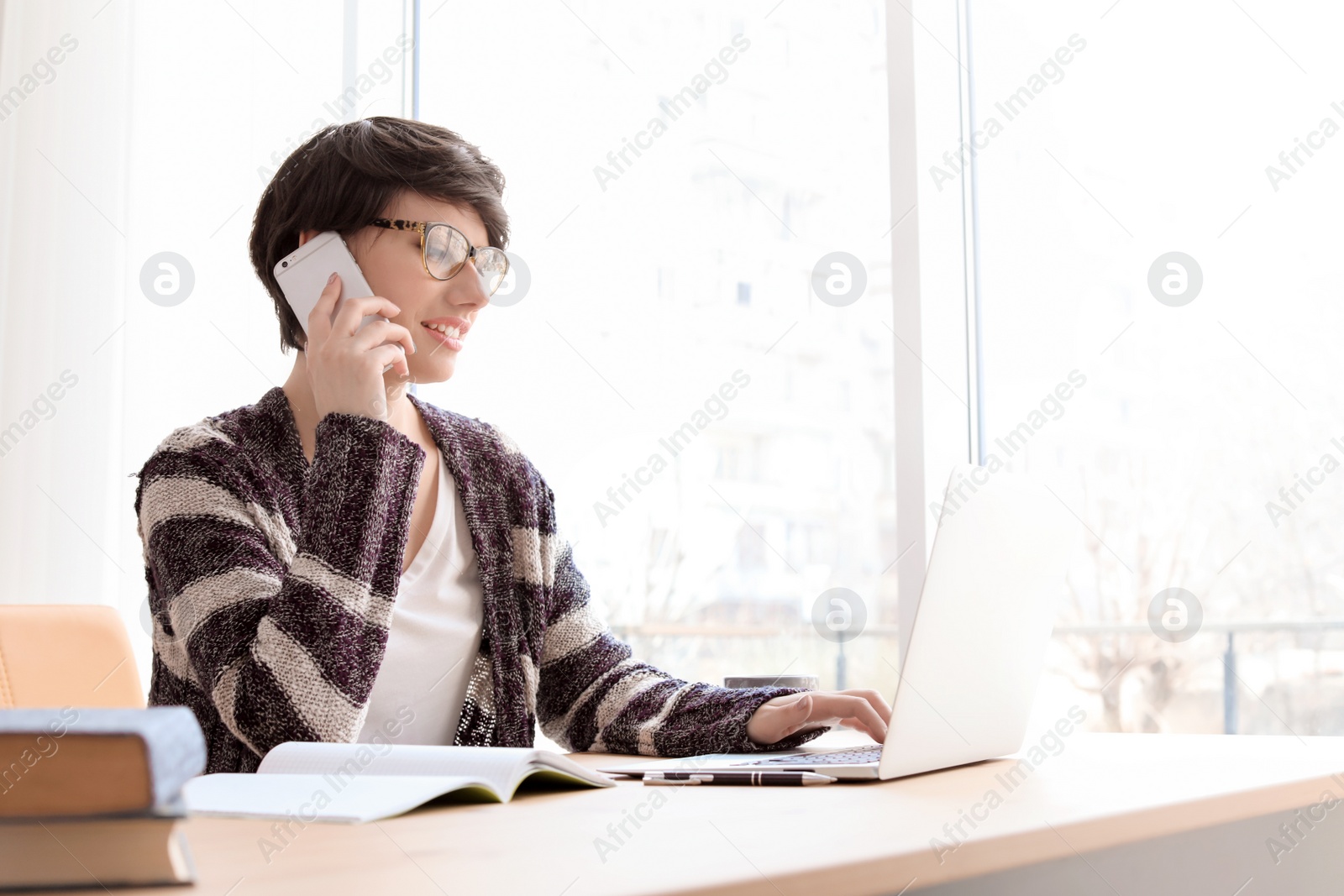 Photo of Young woman talking on mobile phone while working with laptop at desk. Home office