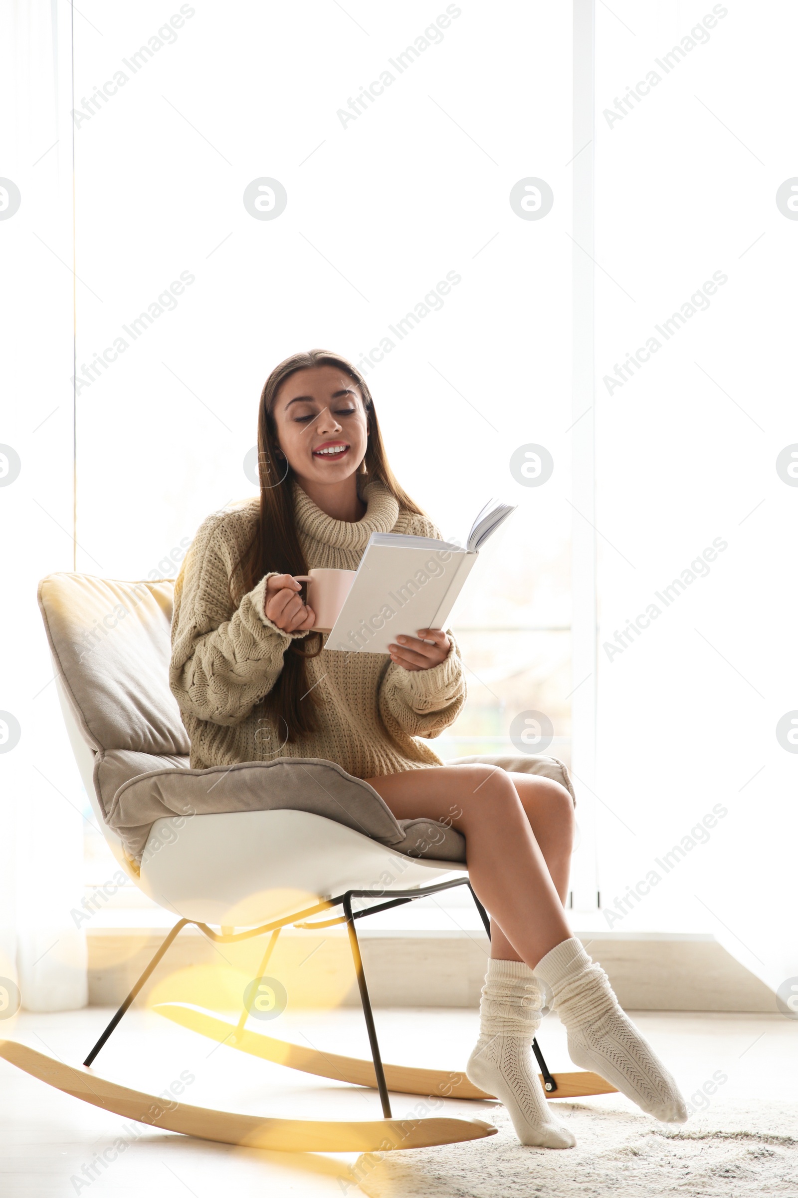 Photo of Young woman with cup of coffee reading book near window at home