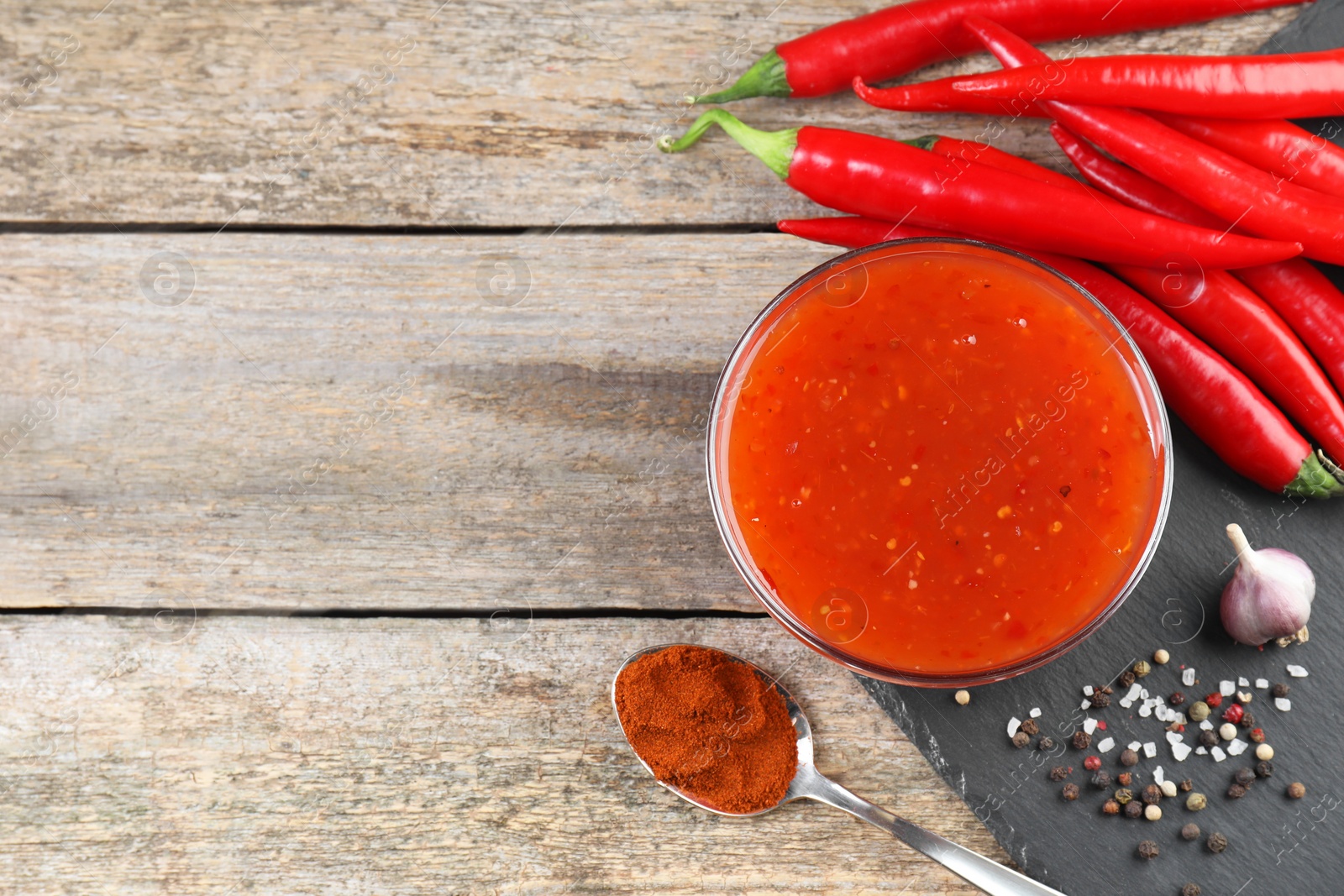 Photo of Spicy chili sauce in bowl and ingredients on wooden table, flat lay. Space for text