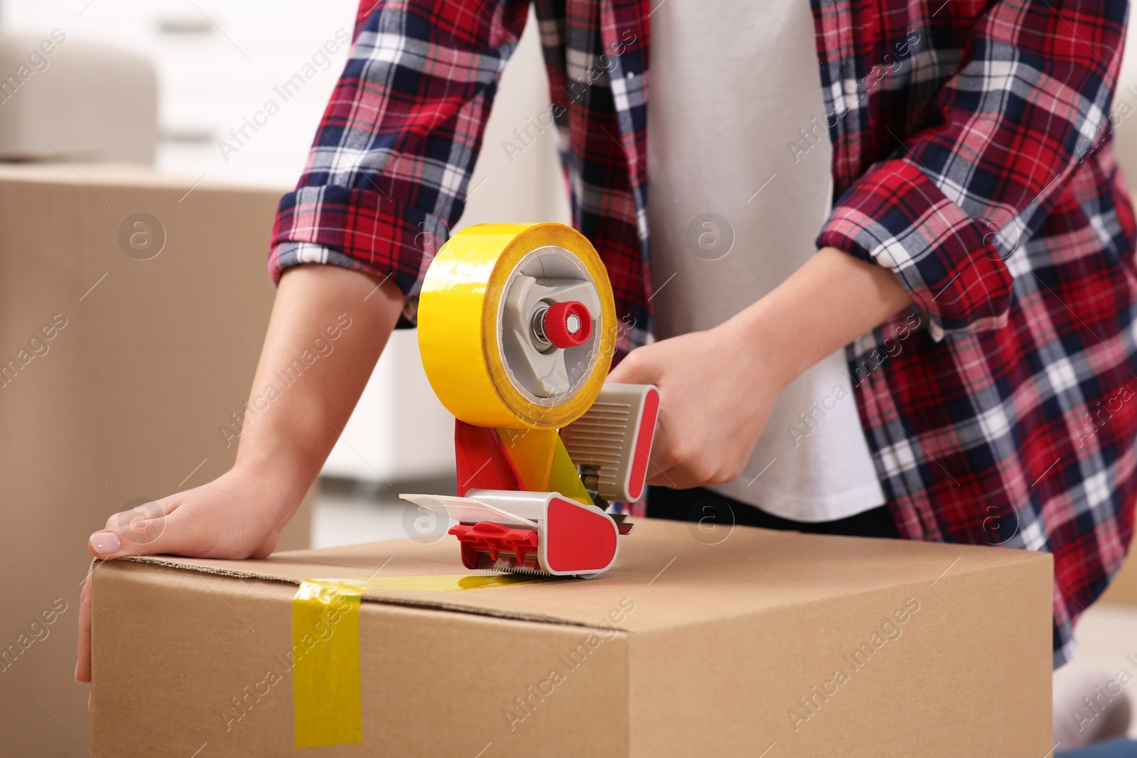 Photo of Woman applying adhesive tape on box with dispenser indoors, closeup