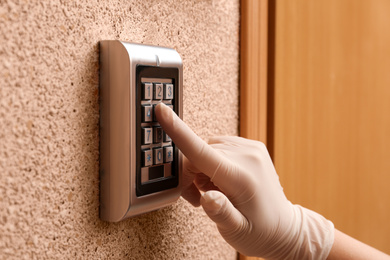 Photo of Woman in gloves entering code on electronic lock's keypad indoors, closeup