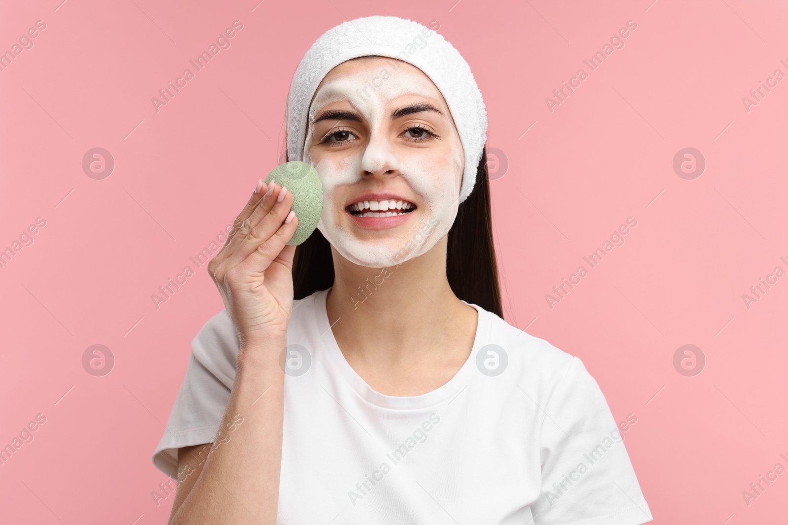 Photo of Young woman with headband washing her face using sponge on pink background