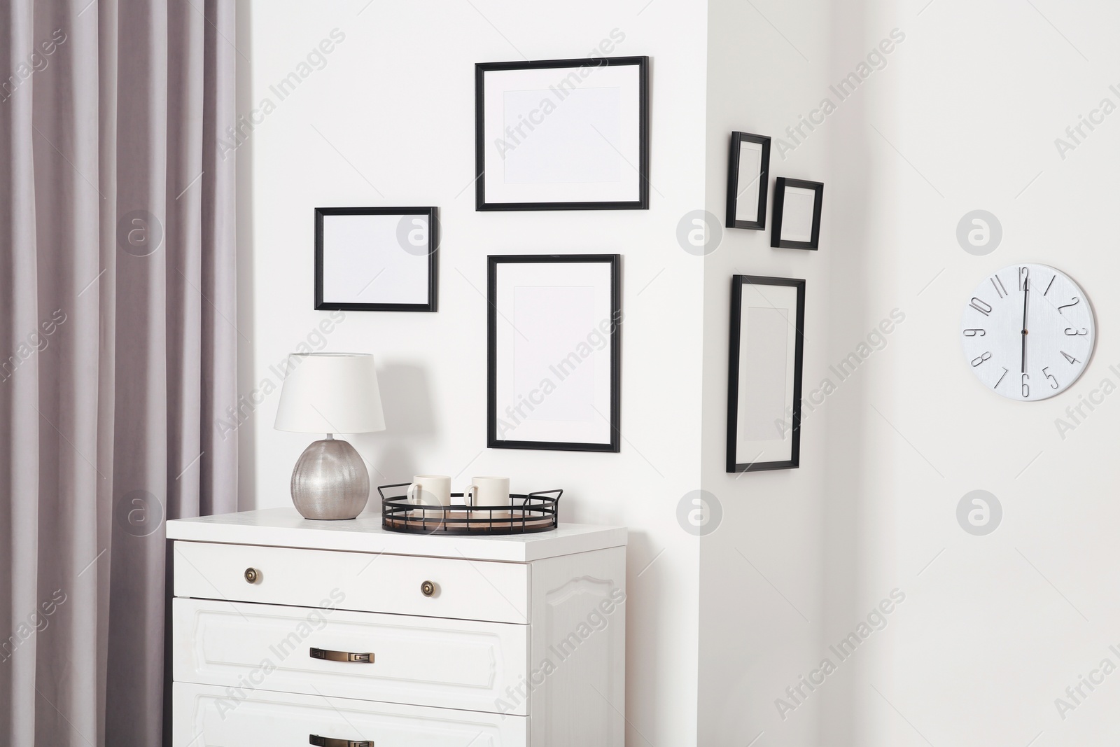 Photo of Empty frames hanging on white wall, wooden chest of drawers and lamp indoors