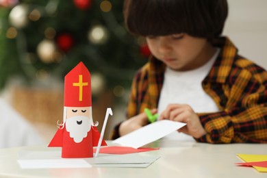 Photo of Cute little boy cutting paper at home, focus on Saint Nicholas toy