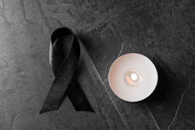 Photo of Black ribbon and burning candle on dark grey stone surface, top view. Funeral symbols
