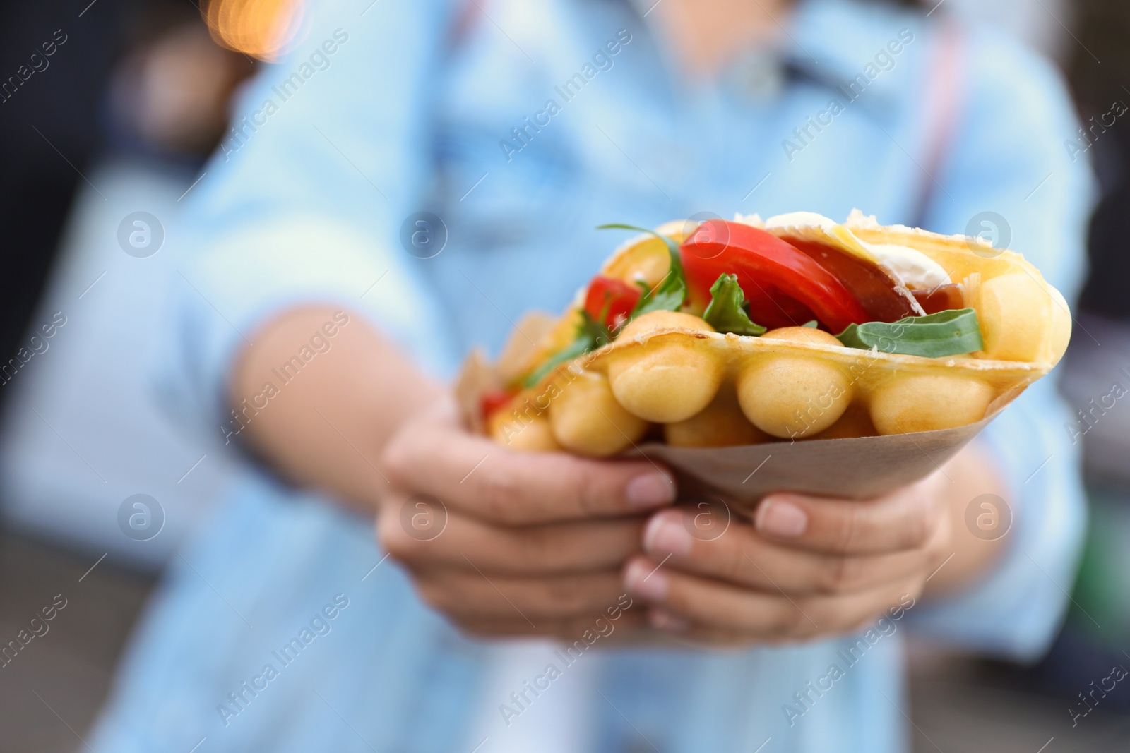 Photo of Young woman holding delicious bubble waffle with tomato and arugula outdoors, closeup