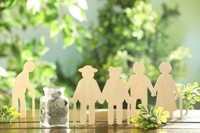 Pension savings. Figures of elderly people, coins in jar and green twigs on wooden table outdoors