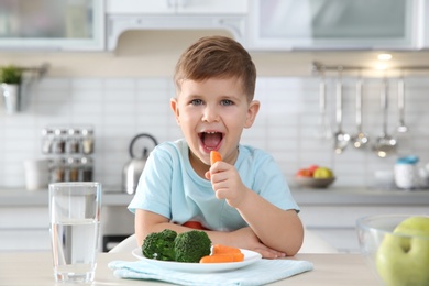 Adorable little boy eating vegetables at table in kitchen