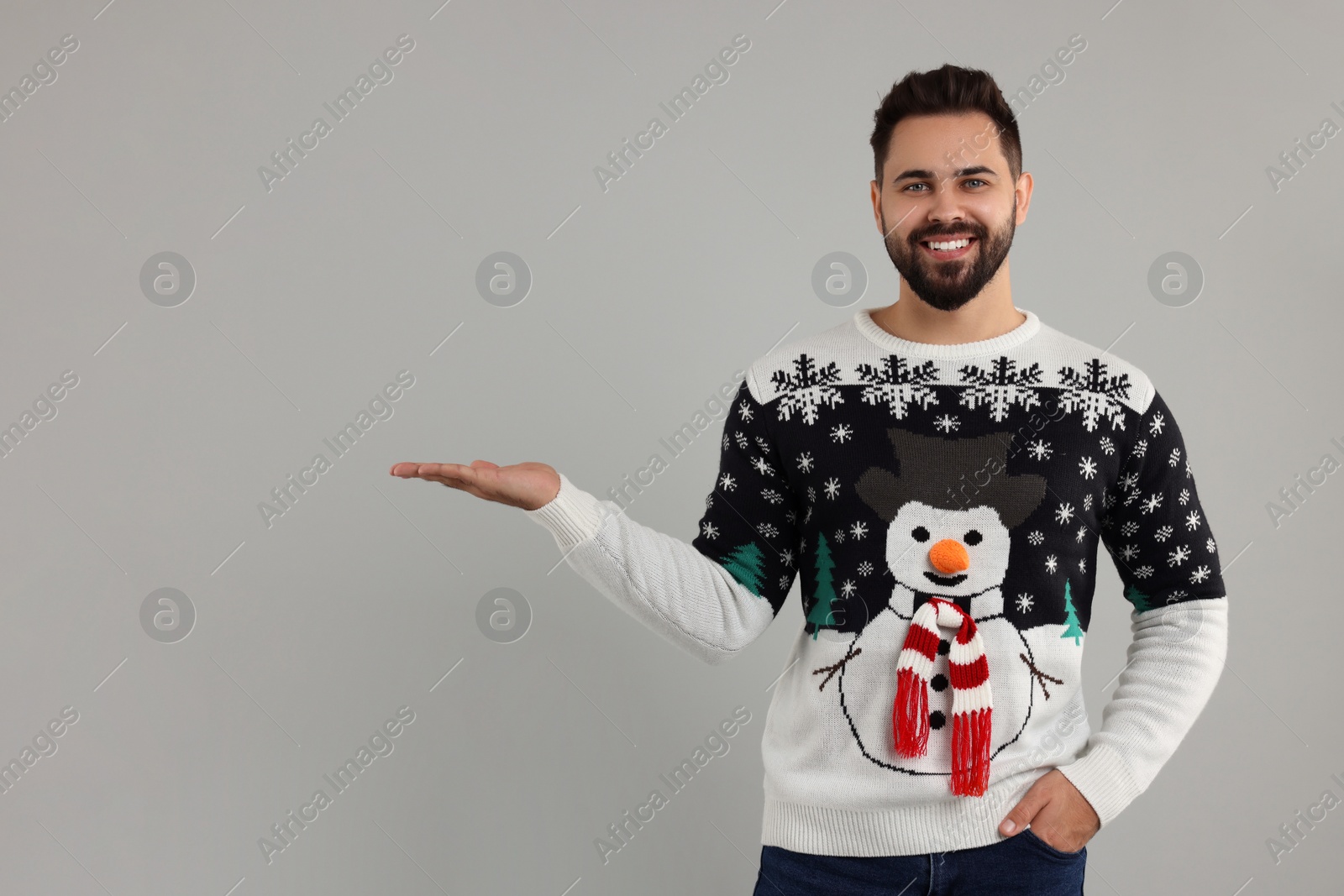 Photo of Happy young man in Christmas sweater showing something on grey background. Space for text