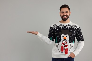 Happy young man in Christmas sweater showing something on grey background. Space for text