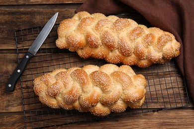 Photo of Homemade braided breads with knife, napkin and cooling rack on wooden table, flat lay. Traditional Shabbat challah