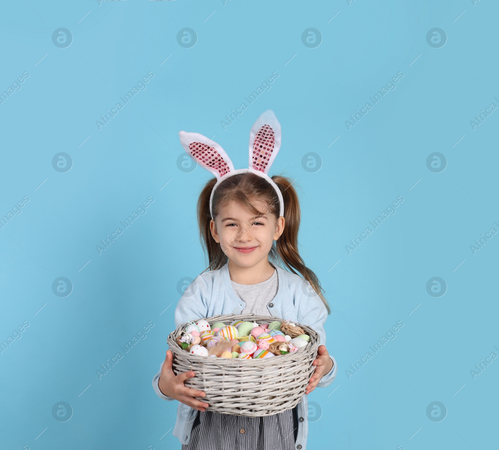 Photo of Happy little girl with bunny ears holding wicker basket full of Easter eggs on light blue background