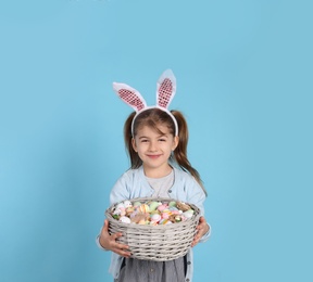 Photo of Happy little girl with bunny ears holding wicker basket full of Easter eggs on light blue background