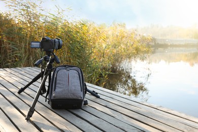 Photo of Tripod with modern camera and backpack on wooden pier near water. Professional photography