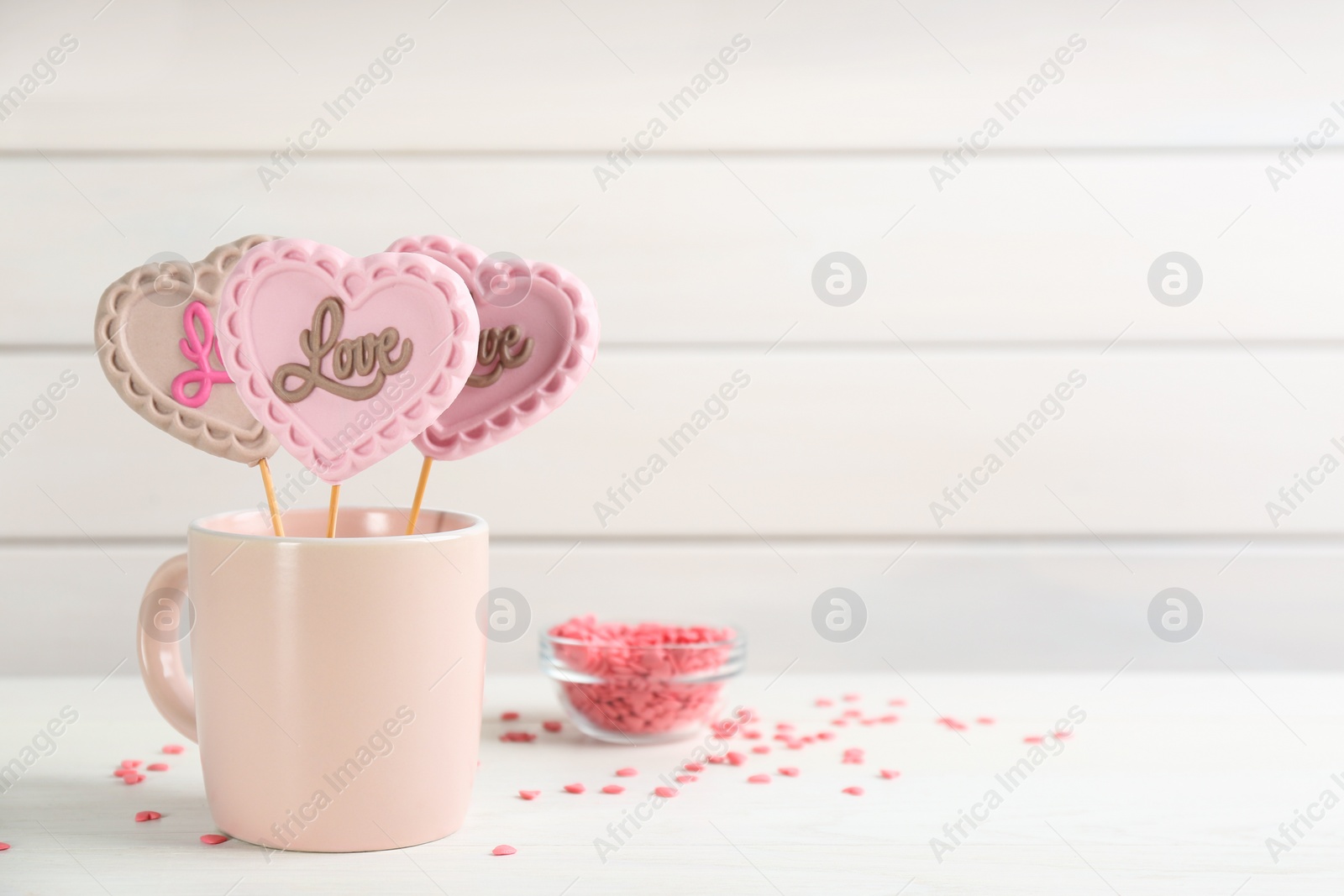 Photo of Heart shaped lollipops made of chocolate in cup and sprinkles on white table, space for text