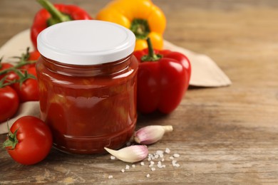 Glass jar of delicious canned lecho and fresh ingredients on wooden table, closeup. Space for text